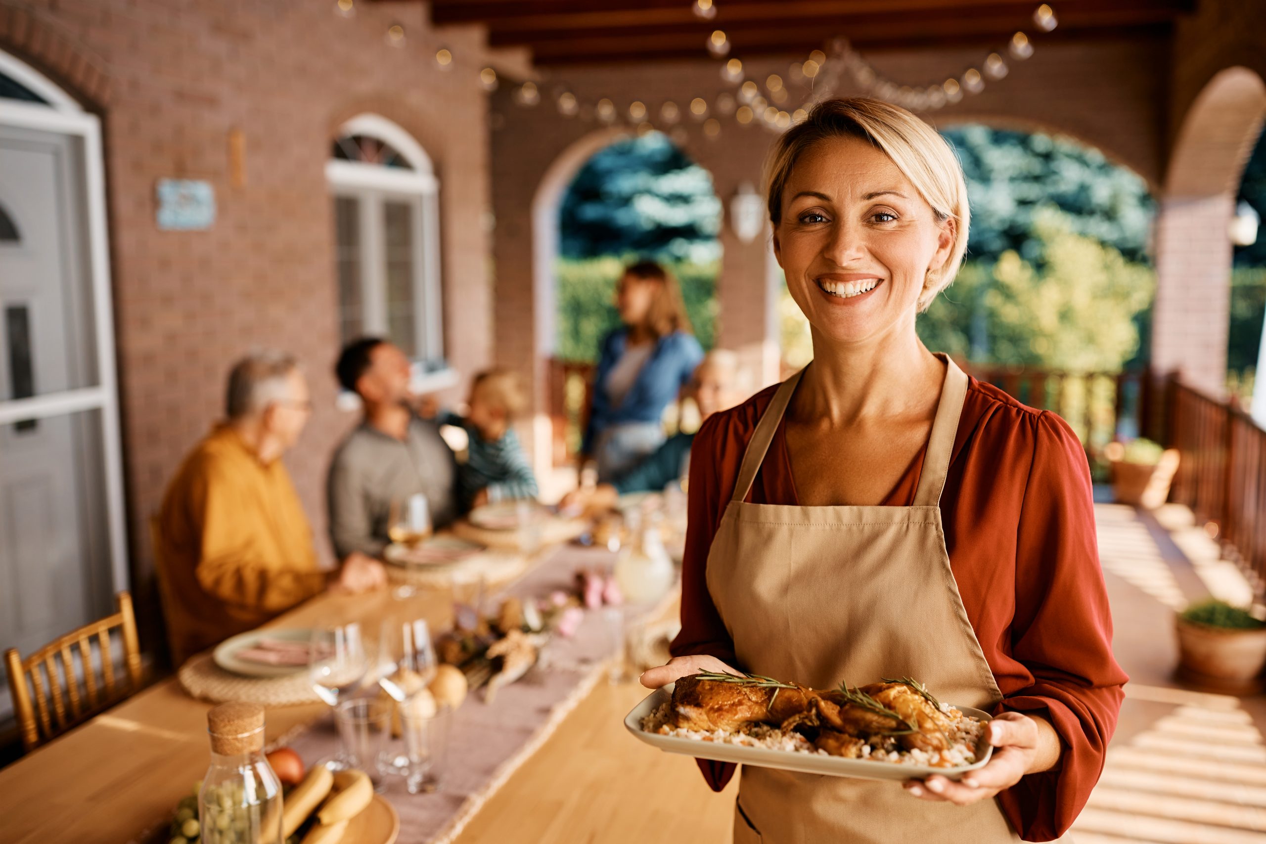 Happy woman serving lunch to her multigeneration family on a terrace and looking at camera.