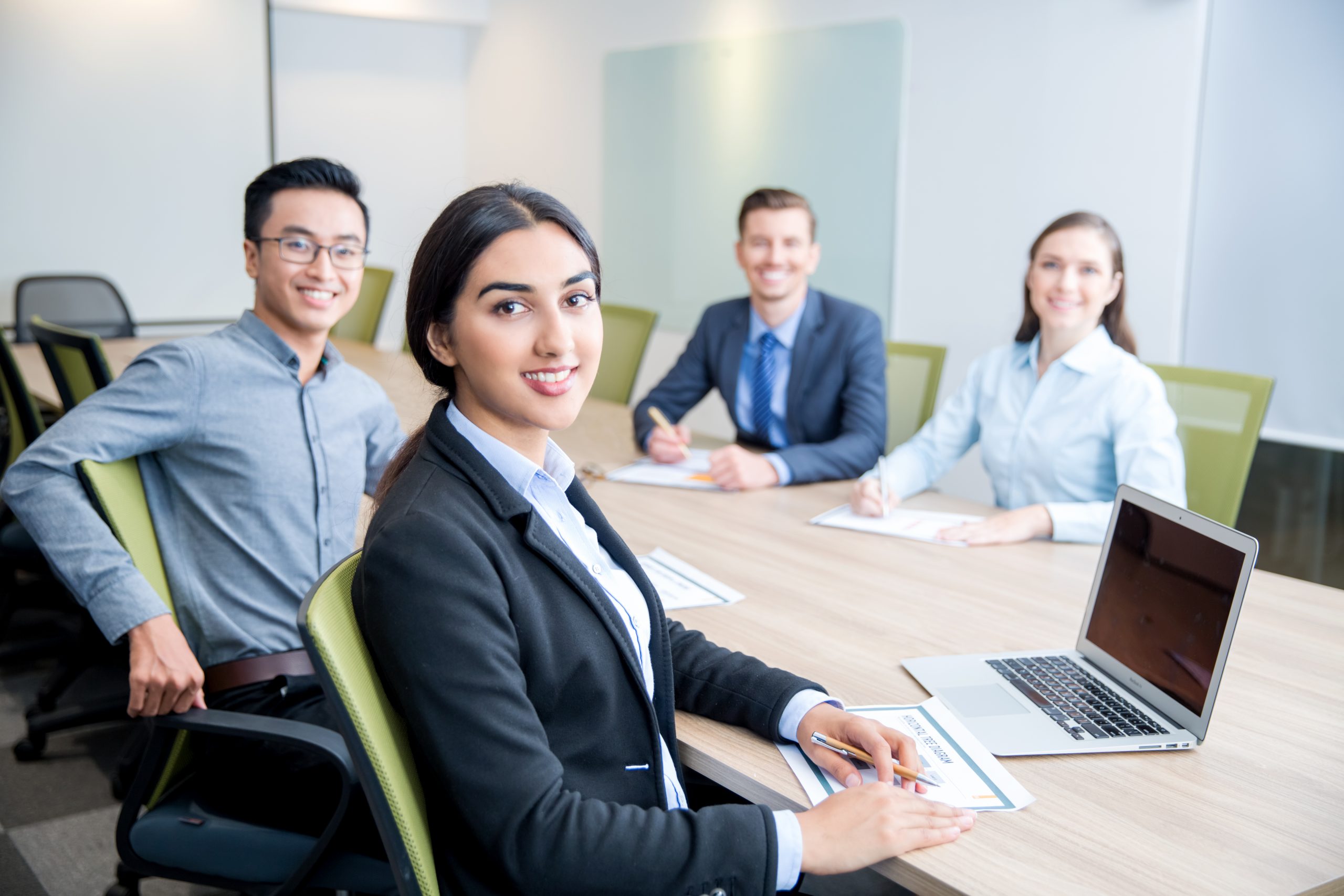 Four smiling middle-aged multi-ethnic business people turning to camera, working and discussing ideas while sitting at big table in conference room. Side view.