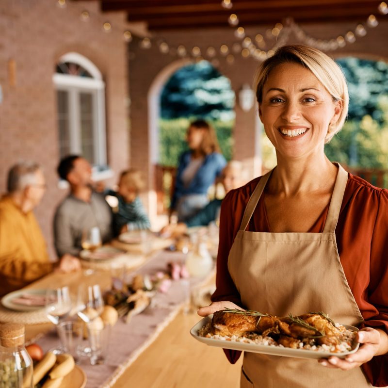 Happy woman serving lunch to her multigeneration family on a terrace and looking at camera.