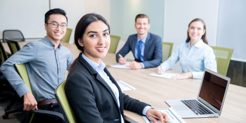 Four smiling middle-aged multi-ethnic business people turning to camera, working and discussing ideas while sitting at big table in conference room. Side view.